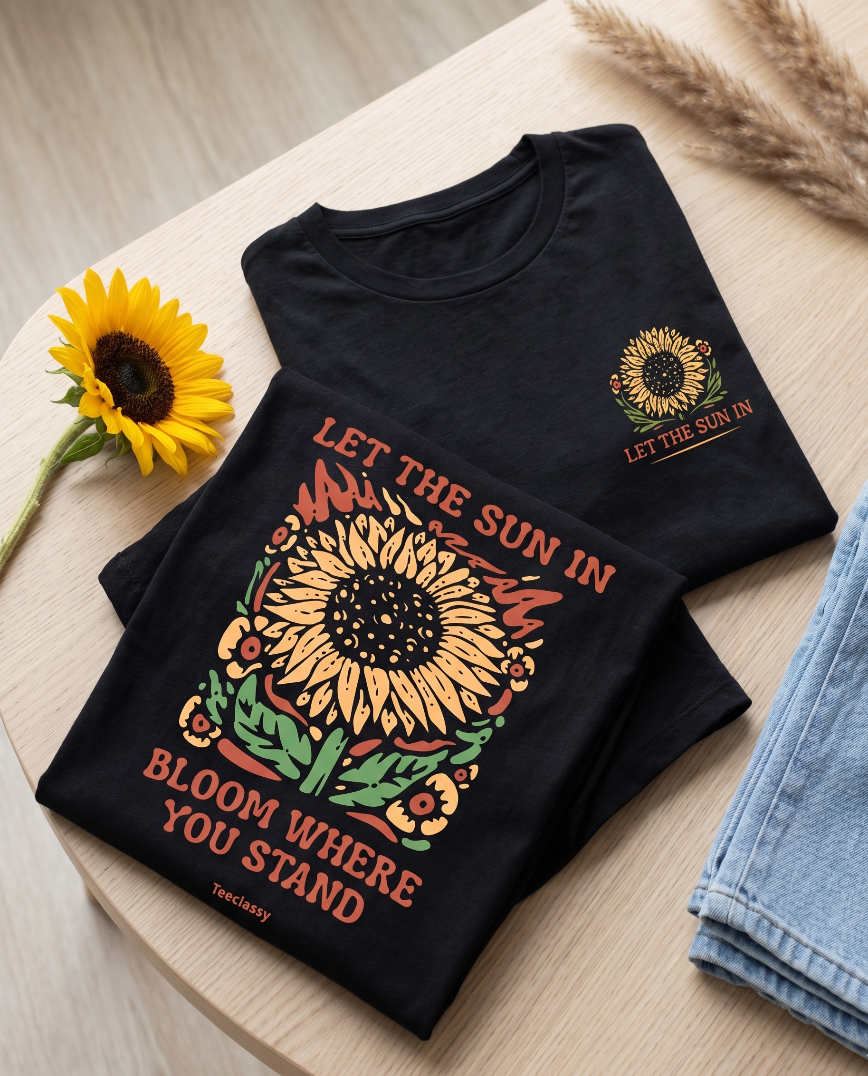 Black t-shirt with sunflower design and text on a wooden surface with a sunflower and denim jeans.